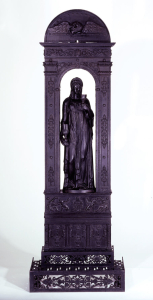 Black cast iron radiator (b) in the form of a podium surmounted by an arch forming a niche for a standing draped figure (a) of a woman- the goddess Hebe- holding a Greek drinking goblet in either hand (d,e). A composite entablature is surmounted by a Doric cornice crowned by a semi-circular tympanum. The podium base is decorated with bas-reliefs of columns alternating with Greek vases surrounded by drapery at the lower level and repeated scene of a griffin and man pouring liquid into a bowl in the upper level. The arch itself is decorated with bas-reliefs of rosettes and scrolls on two supporting pilasters with fluted capitals. The tympanum has a bas-relief of an eagle clutching a staff from which springs ribbons bearing "Stratton" and Seymour". Stars decorate tympanum, following the semi-circular curve. Radiator stands on four detachable scrolled legs (f/i). The fender (c) is comprised of grille work formed by scrolls, acanthus leaves and rosettes. Flat circular flue key (j) with stylized foliate handle fits on pipe behind tympanum.