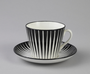 Straight-sided, tapering circular cup (a) with glazed decoration consisting of series of narrow black vertical wedges on white ground; black along edge of rim; white loop handle and white interior. Circular saucer (b) with upraised rim and same tapering black decoration on white ground; black at rim; white underside.