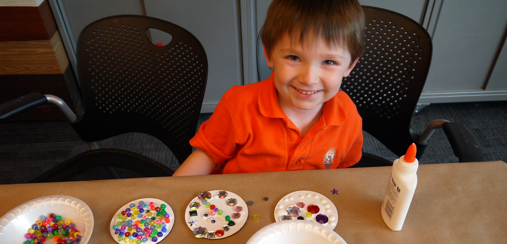 boy smiles and looks at camera, with glue bottle and buttons on a table in front of him