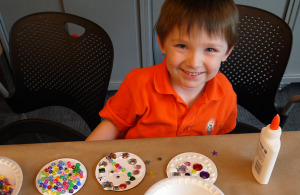 boy smiles and looks at camera, with glue bottle and buttons on a table in front of him
