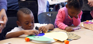 Two children working concentratedly with colored paper and clue and crayons.