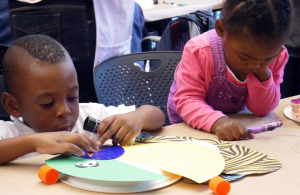 Two children working concentratedly with colored paper and clue and crayons.