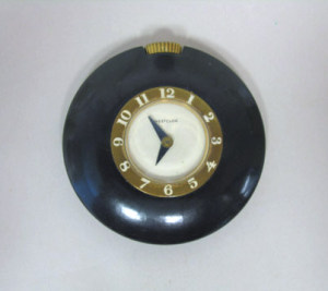 Circular form; smooth black case surrounding metal band with white numerals encircling white face with black hands; metal stem at top.