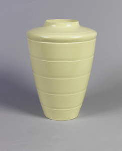 Tapering cylindrical form with circular mouth, broad shoulder; body decorated with horizontal grooves; creamy yellow matte glaze.