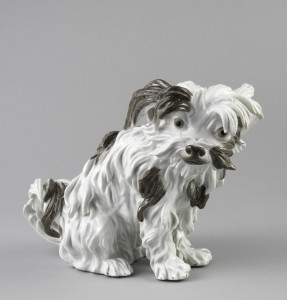 Figure of small long-haired dog, sitting, head slightly turned; glazed in white with brown patches.
