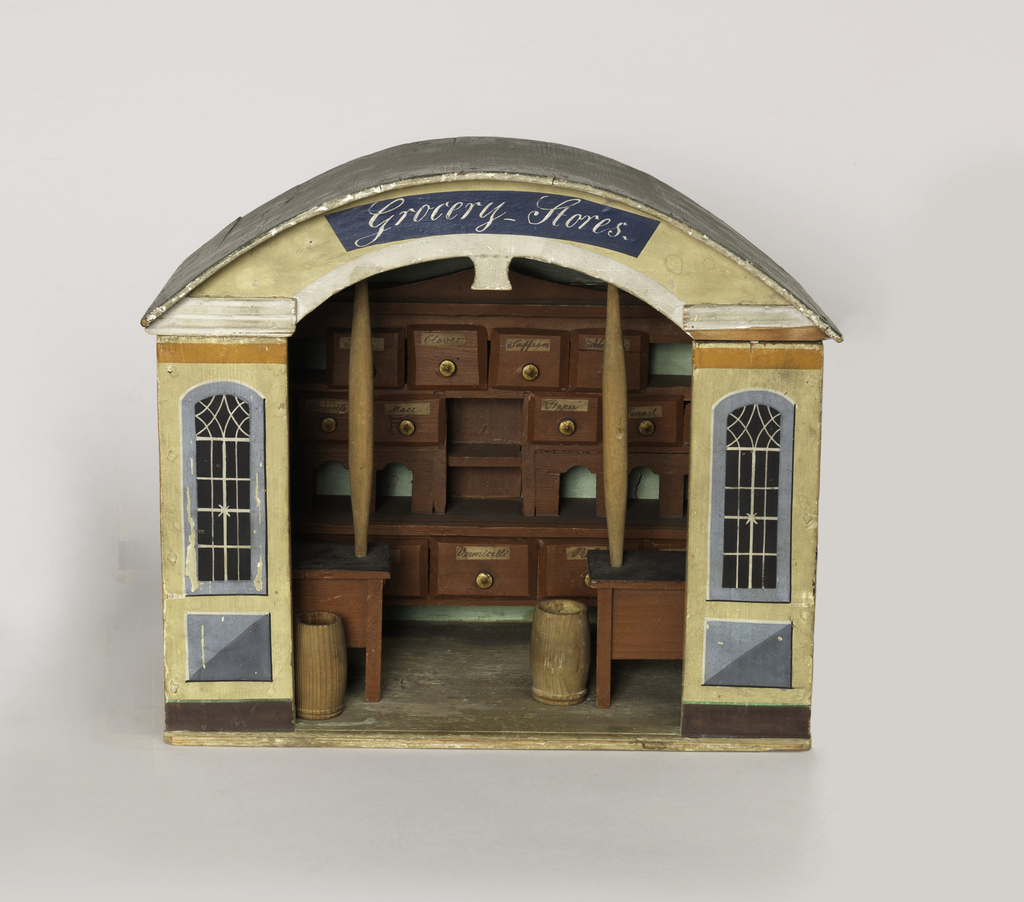 Miniature of a store made with solid back and two sidewalls, front panels showing two painted windows and central opening for doorway with "Grocery Stores" above it. Against the back wall, a cabinet with shelves and partitions and 16 drawers (-2b - -2l) each with a gold painted knob and handwritten paper label. One each for: almonds, annis seed, cacao, cinnamon, cloves, fenel, mace, millet, pepper, pimento, raisins, rice, saffron, sape, vermicelle (one drawer not labelled); two free-standing barrels (-2l and -2m), and two tables (-2n and -2o), each with a post (-2p and -2q) supporting one end of an arch (-2r) from which hangs a pair of scales (-2x - -2z). Pencil scribblings over outside of structure.Possibly English. In 1820's style, possibly early 19th century.