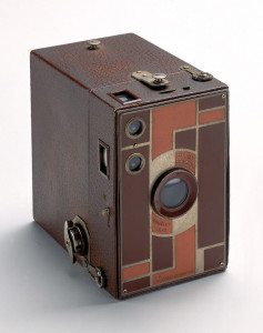 Box camera (a) covered in brown leatherette; metal faceplate enameled with tan and brown geometric design. Camera slides into box shaped leatherette-covered case (b).