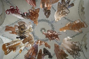 A photograph taken from the ground looking up at various brown, white, and orange garments hanging from an ornate ceiling.