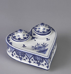 Heart shaped form, horizontally placed on three small feet. Circular openings in two lobes, with two inkwells, each with cover. Slight depression on top, at point. White ground with blue decoration of stylized flowers and foliage.