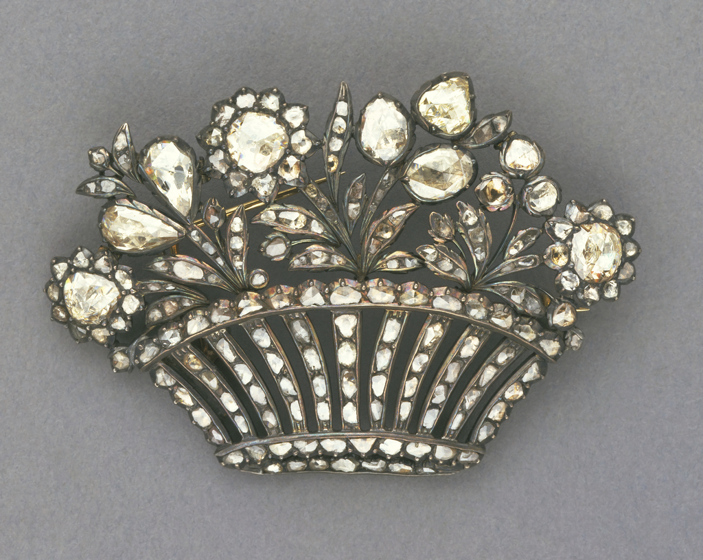 In the shape of a low, wide basket filled with a floral and foliate arrangement, composed of rose-cut diamonds set in silver and gold. Reverse features closed settings; double-pronged pin, the longer of which slips under c-clasp. Possibly originally part of a tiara or a hair ornament.