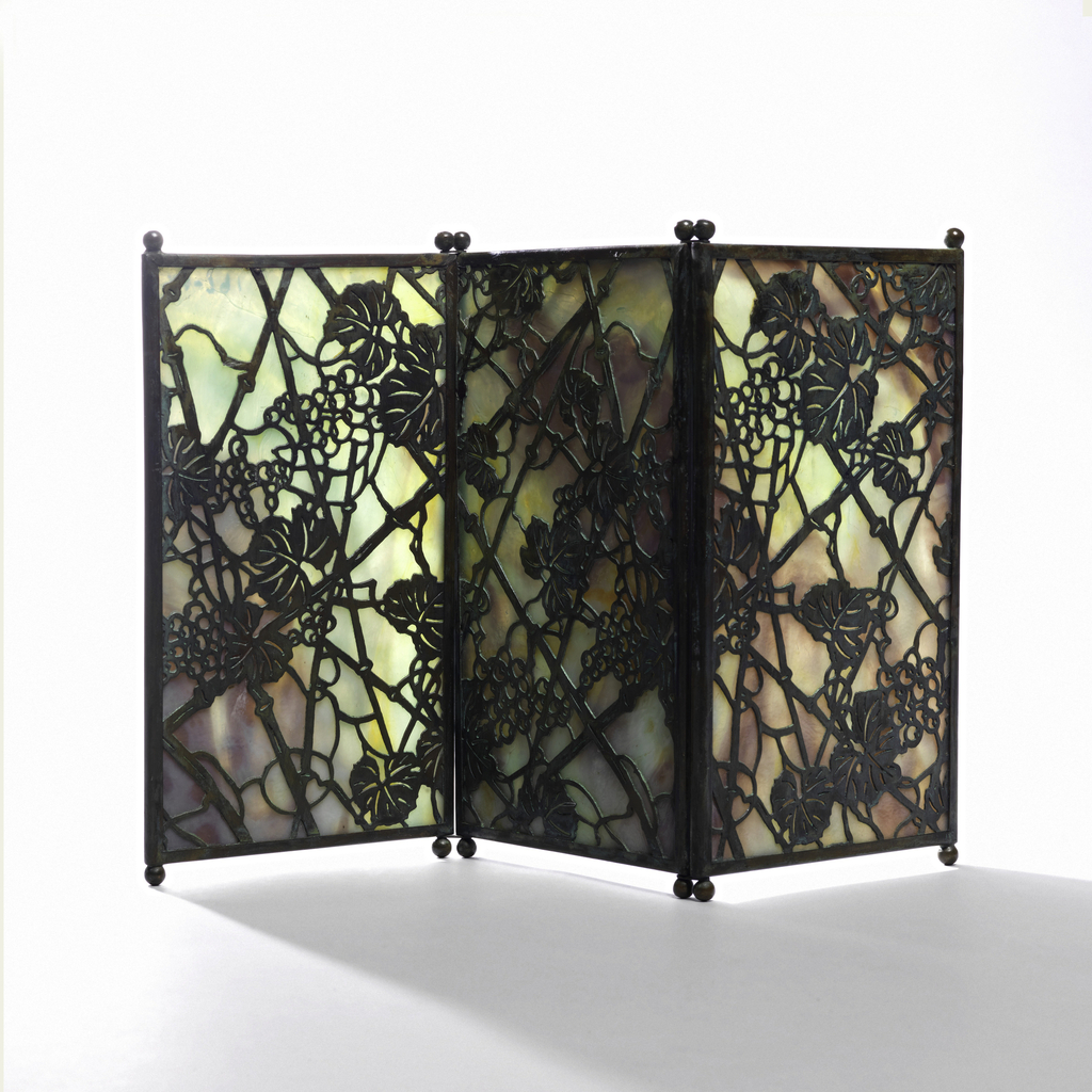 Three vertically rectangular panels of marbleized glass, each panel framed with narrow copper edging oxidized to green and orange-red tones with two small ball feet at each end. The panels are hinged together by means of wire at juncture of feet and screen. Cut out copper overlay. Glass marbleized in tones of lavender, blue and green overlaid with cut out copper in “Grapevine” pattern.