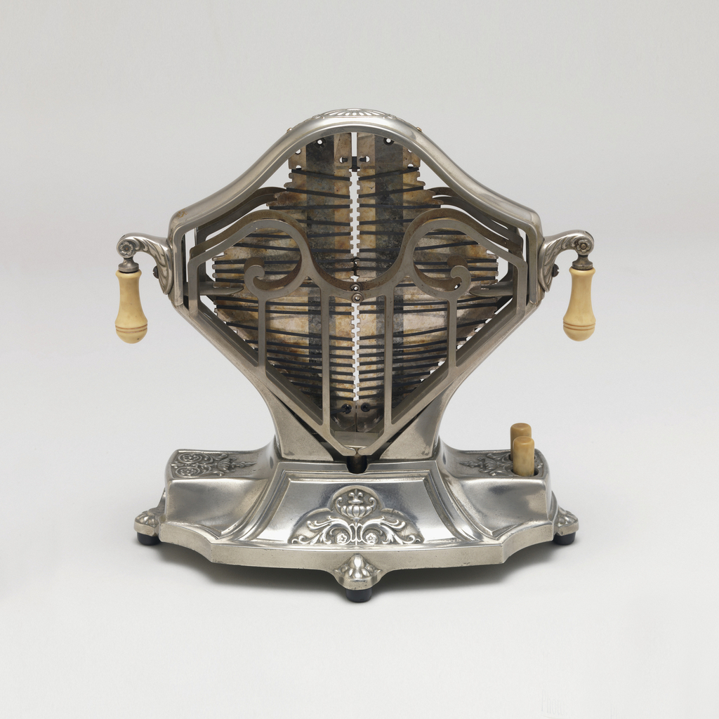 An early metal toaster in a diamond shape with a visible heating elements and metal frames to hold the toast on either side. Roughly rectangular cast metal base with small decorative floral designs near the feet; two cylindrical buttons on one side. Small pendant knobs on opposite sides of toaster.