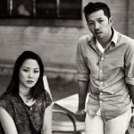 A black and white photograph of Carol Lim and Humberto Leon. Lim sits in a chair and Leon leans against a table beside her.