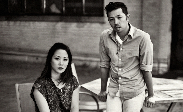 A black and white photograph of Carol Lim and Humberto Leon. Lim sits in a chair and Leon leans against a table beside her.