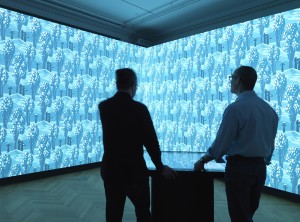 Two silhouetted figures stand in front of an interactive multi-touch table inside of the Immersion Room. The design which they are creating through the touch table drawing application is projected on two walls in front of them.