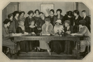 a group of fashionable people sit and stand around a table