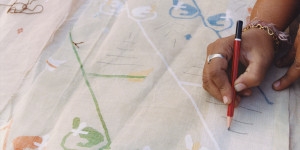 An artisan's hands hold a pencil and draw a series of short lines on woven fabric to mark where appliqué designs will be applied.