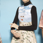 Three models are photographed from waist to neck standing beside each other. The model on the left is wearing a white collared shirt under a sweater with an illustration of sand and blue skies with two floating disembodied eyes and a mouth. The middle model wears a black turtleneck with a dress over it. The dress has a similar beach scene with eyes and a mouth. The model on the right wears a t-shirt with a brown to pink gradient sunset sky with a disembodied eye, nose, and mouth.