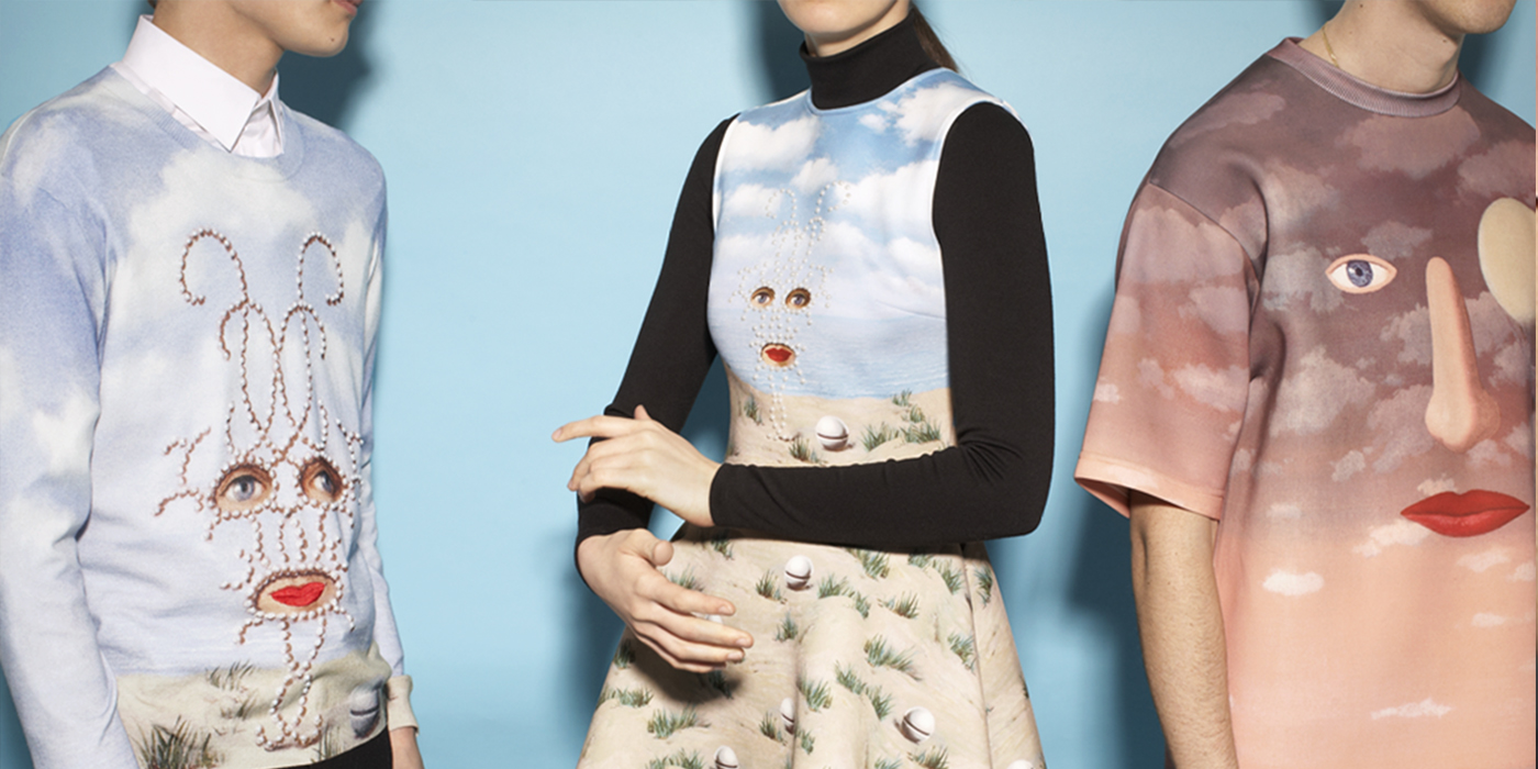 Three models are photographed from waist to neck standing beside each other. The model on the left is wearing a white collared shirt under a sweater with an illustration of sand and blue skies with two floating disembodied eyes and a mouth. The middle model wears a black turtleneck with a dress over it. The dress has a similar beach scene with eyes and a mouth. The model on the right wears a t-shirt with a brown to pink gradient sunset sky with a disembodied eye, nose, and mouth.