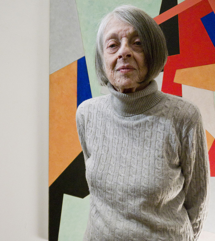 Elaine Lustig, an older woman with light skin and gray chin-length hair, stands in front of a blocky, abstract, colorful wall and looks serenely at the camera with her hands clasped behind her back.