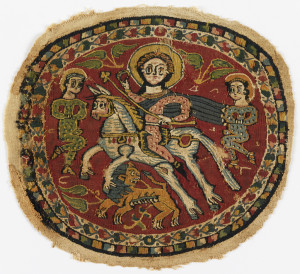 Tapestry medallion with polychrome figures on a red ground. In the center field, a haloed rider on a prancing horse carries a staff or scepter topped with a trefoil, and holds a ring. A lion below looks back over his shoulder. Two additional figures, one at each side, wear spotted green clothing. The rider is in pink with a flowing blue cape. Inner border of polychrome connected flower heads on a natural ground; outer border of polychrome stepped pattern on dark blue ground. The image is said to represent the emperor with two Persian prisoners, a reference to the victory of Heraclius (617 - 641) over the Persians in 627.