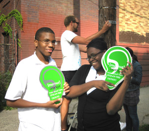 two young people hold green paper "ears" while man installs a sign on a telephone pole