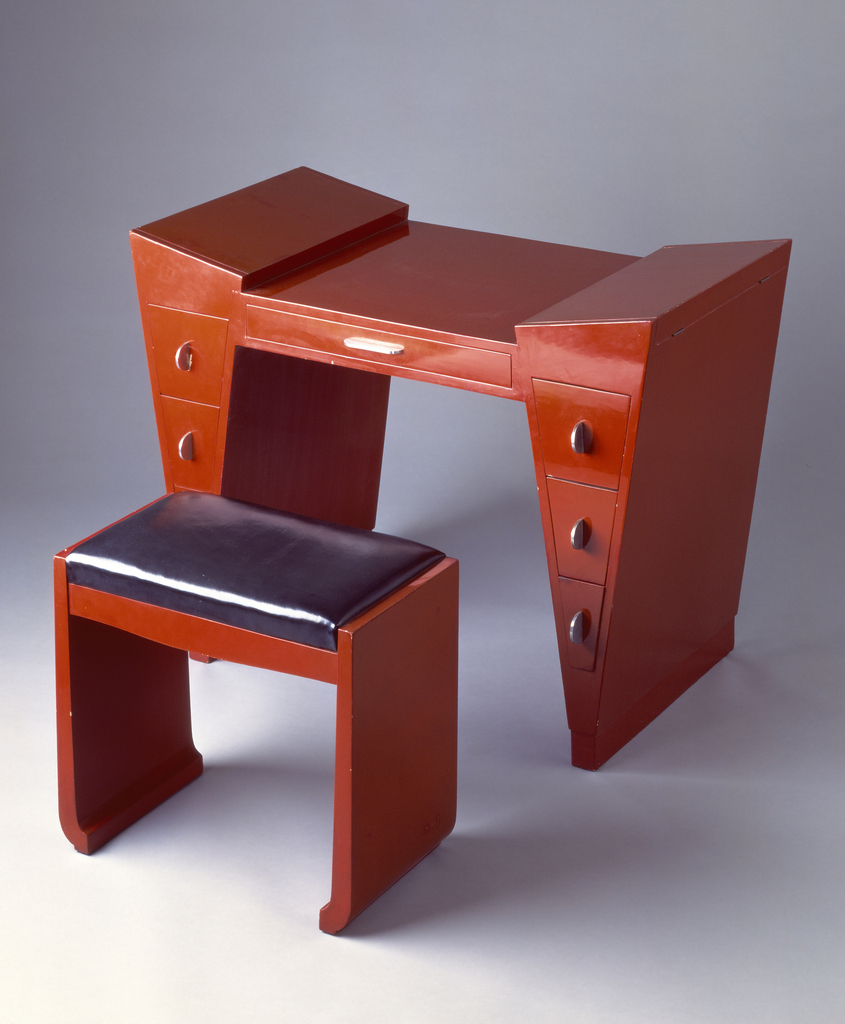 Red-lacquered wood dressing table and bench of geometric form; dressing table with hinged, covered storage wells at top of each tapering leg, and three drawers below, each with semi-circular metal pull; one wide drawer in center with horizontal metal tab pull; bench with rectangular black leatherette seat cushion.