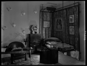 interior view of a study designed by Pierre Chareau at Lord & Taylor in 1928. The interior consists of a desk with a chair, a collapsible side table, a suspended bed/seating area, and a wall adorned with a geometric-patterned curtain, four works of art, and a number of geometrically styled sconces
