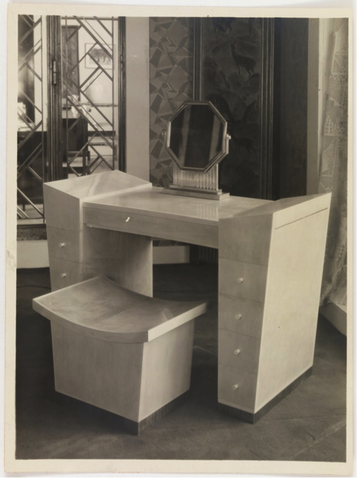Photograph of an angular dressing table topped by an octagonal mirror. The table is in situ of a complementary modern interior, with numerous geometric pattens on surfaces behind it, including a mirrored cabinet and a wallcovering or wall hanging.