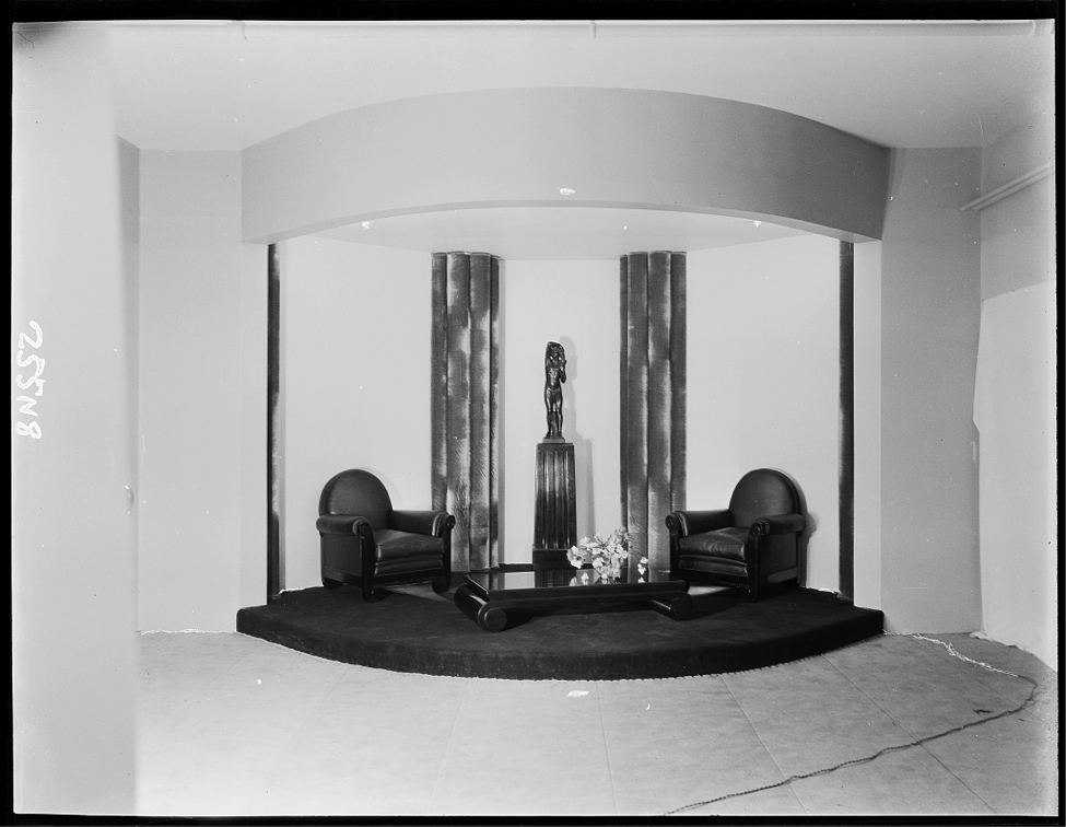 View of a liviing room designed by Sue et Mare at Lord & Taylor. The round display consists of two padded arm chairs, a low coffee table with rounded legs, and a tall, paneled plinth on which stands a statue of a nude figure.