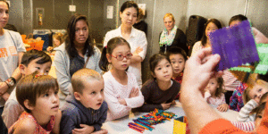 Many youths look on as a person holds up a popsicle stick construction