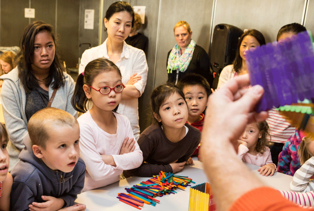 Many youths look on as a person holds up a popsicle stick construction