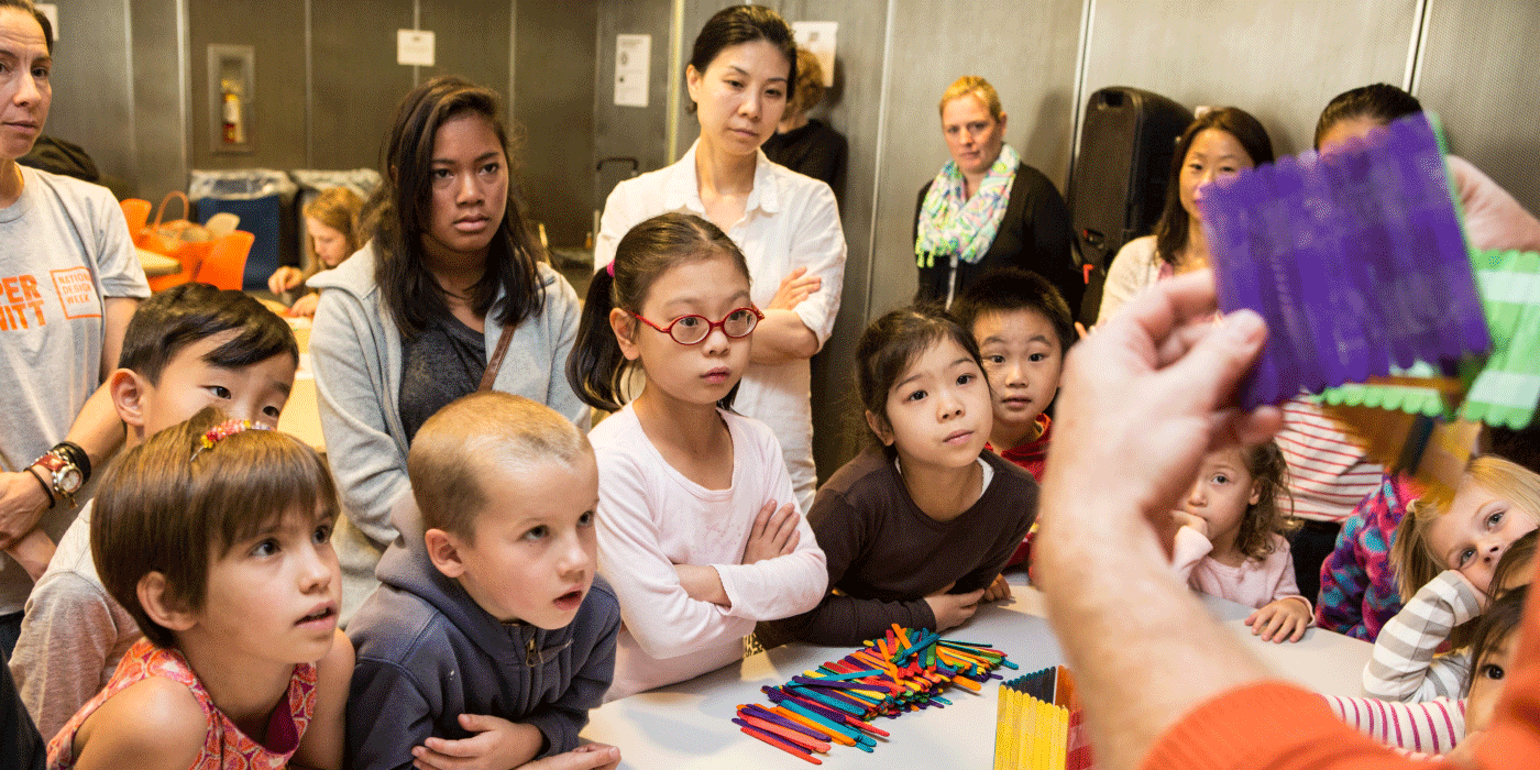 Many youths look on as a person holds up a popsicle stick construction