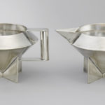Two silver sugar bowls shaped vaguely like UFOs or gemstones, with a wide, round opening that tapers to a bottom point that rests on four triangular feet. The bowl on the left has two square handles, and the bowl on the right has one square handle and one spout.