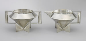 Two silver sugar bowls shaped vaguely like UFOs or gemstones, with a wide, round opening that tapers to a bottom point that rests on four triangular feet. The bowl on the left has two square handles, and the bowl on the right has one square handle and one spout.