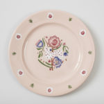 Light pink circular plate with a stylized design in the center in red, green, blue, and white featuring a polka dot deer leaping in front of several flowers.