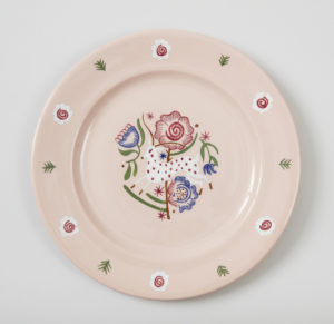 Light pink circular plate with a stylized design in the center in red, green, blue, and white featuring a polka dot deer leaping in front of several flowers.