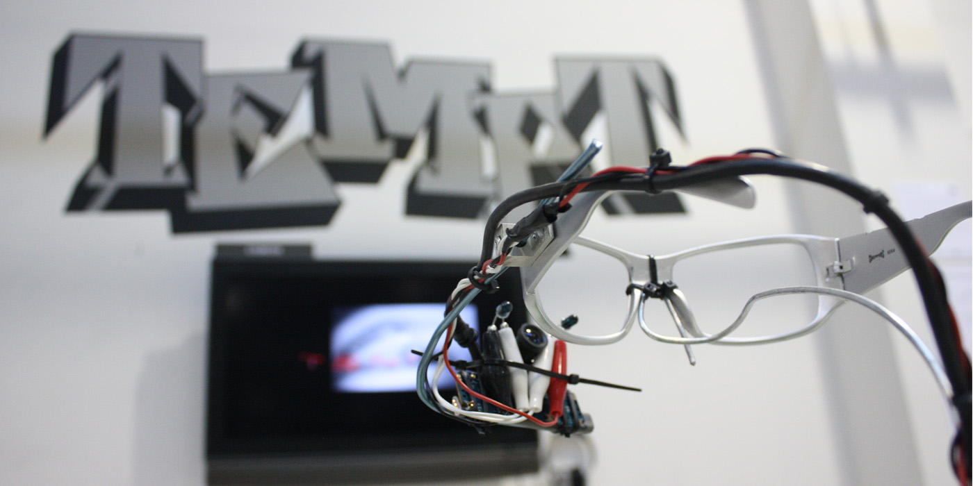 Photograph of white eyeglasses connected to wires. The glasses face a screen and a wall that says "TEMPT."