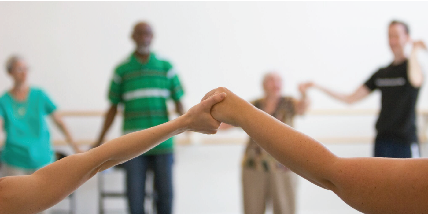 Photograph of six people in a dance studio. The photograph focuses on two people's clasped hands. Four people stand in the background holding hands.