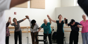 Photograph shows seven people in a dance studio. All hold hands with their arms lifted up.