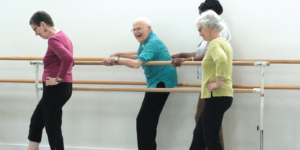 Three people with legs in air dance holding on to ballet bars at waist and shoulder height.