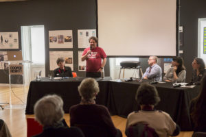 Image of Paul Orselli, President and Chief Instigator, POW! and blogger ExhibiTricks, standing to introduce himself during a panel discussion at Cooper Hewitt. Audience members and panelists look on. Paul wears a red t-shirt with the words "Museums are not neutral" printed on it.