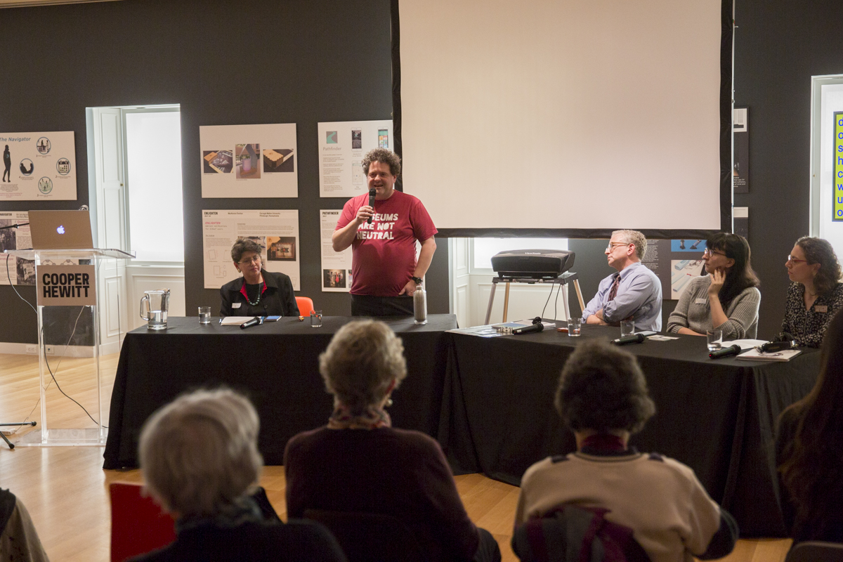 Image of Paul Orselli, President and Chief Instigator, POW! and blogger ExhibiTricks, standing to introduce himself during a panel discussion at Cooper Hewitt. Audience members and panelists look on. Paul wears a red t-shirt with the words "Museums are not neutral" printed on it.
