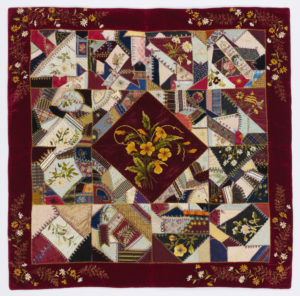 Image features: Patchwork cover made from a variety of woven fabrics and ribbons. Diamond shape in the center with a triangle on each side to form a central square surrounded by twelve squares plus an outer border. Each square contains an embroidered naturalistic flower. Please scroll down to read the blog post about this object.”