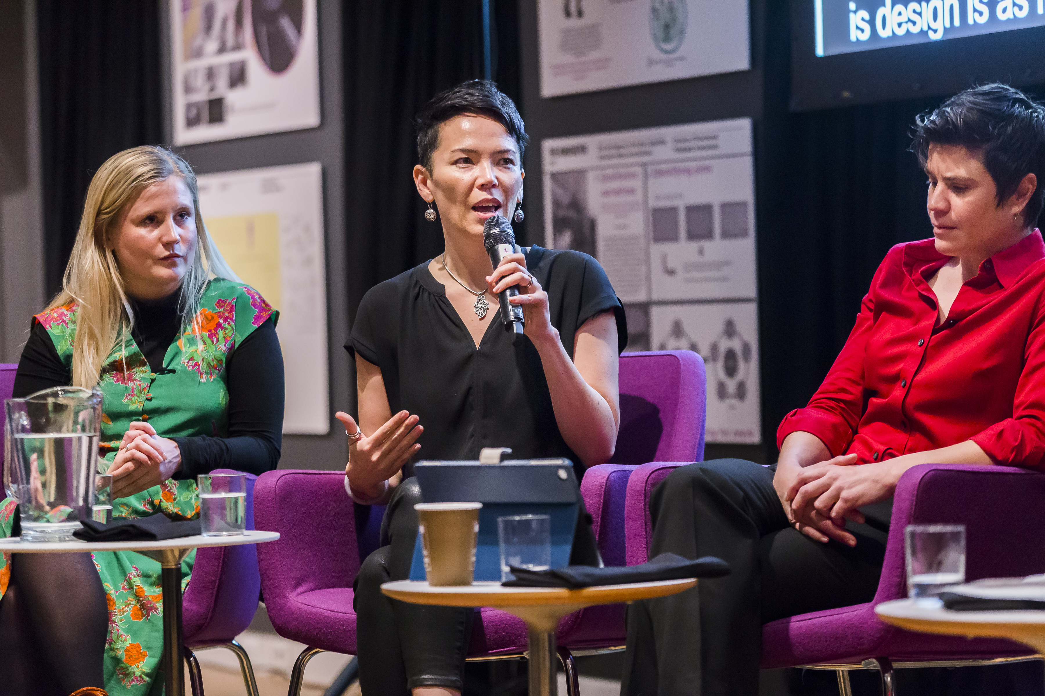 Image of three women sitting in purple chairs during a panel discussion. The woman in the middle Kat Holmes, speaks into a microphone