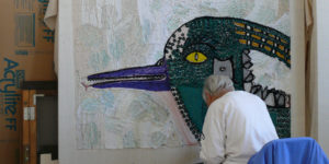 Photograph of Creative Growth studio featuring a large canvas with a n incomplete rug on it. A seated man works on the rug.