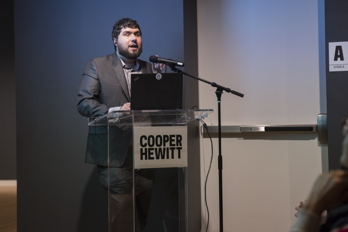 Image of Sina Bahram,Director of Prime Access Consulting (PAC), at a podium, giving a lecture to a group of 50 people sitting around tables.