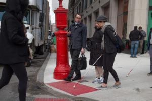 A person uses a white cane to determine the corner of a sidewalk. Click here to listen to a video of the museum's Designing Accessible Symposium.