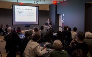 Image of Sina Bahram,Director of Prime Access Consulting (PAC), at a podium, giving a lecture to a group of 50 people sitting around tables. There is a projector screen on the left side of the image with a bright white text slide that reads "Ways to think about disability"