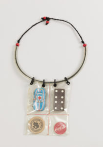 Necklace with pendant suspended from four coiled plastic loops on clear acrylic tube strung with black hemp cord knotted with four red plastic pony beads. Pendant: clear vinyl pouch stitched with red thread to form four sections,each holding a found object: a die-stamped tin toy car; a wooden domino tile; a wooden nickel; and a small wooden disk showing the Coca-Cola logo. Please scroll down to read the blog post about this object.